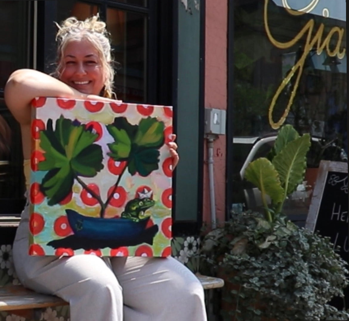 A blonde girl  dressed in white striped overalls and a yellow tube top smiling while holding her artwork of a frog in a boat with large green foliage and red circles in the background, sitting outside the frong stoop of Cantina Gia in Ottawa, Ontario.