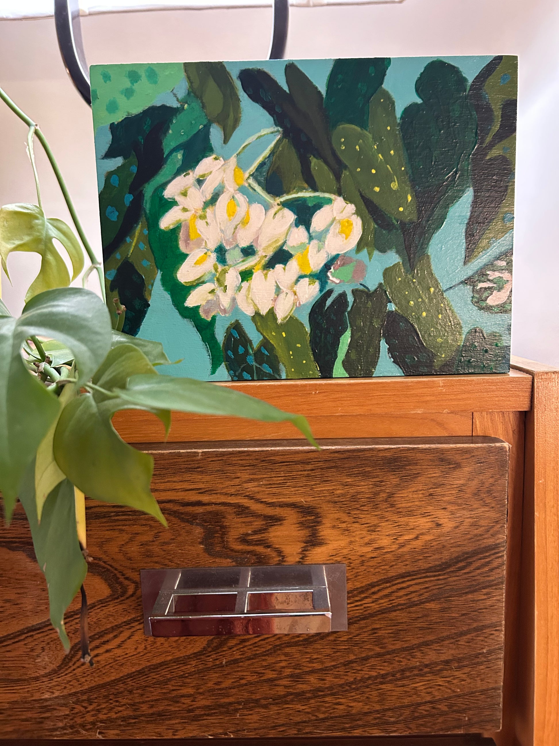A painting of white flowers and green leaves on a blue background sitting atop of a dresser with some green leaves coming through the left hand side of the photograph. 