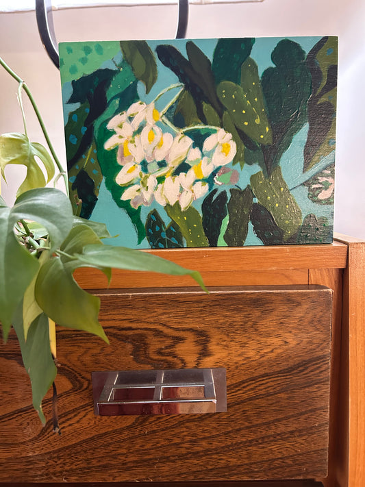 A painting of white flowers and green leaves on a blue background sitting atop of a dresser with some green leaves coming through the left hand side of the photograph. 